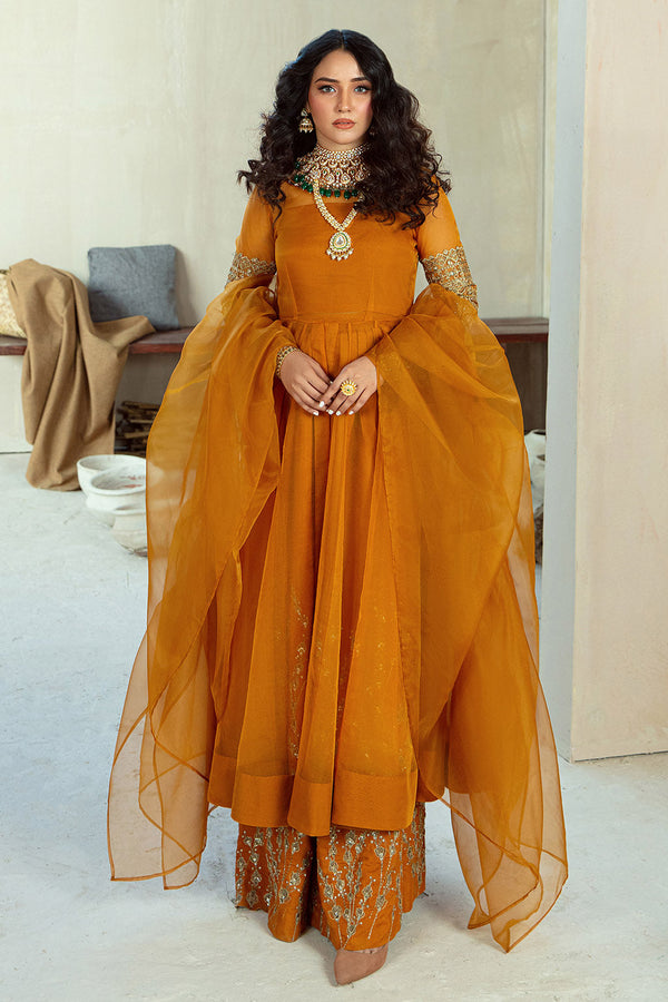 A woman wearing a mustard yellow organza and raw silk frock with embroidered sleeves and dupatta, standing in a room with a plain background.